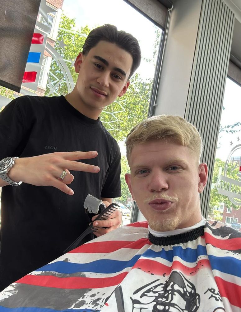 Barber posing with clippers beside a client with a short blond crop